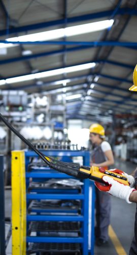 Factory worker wearing uniform and hardhat operating industrial machine with push button joystick in production hall.